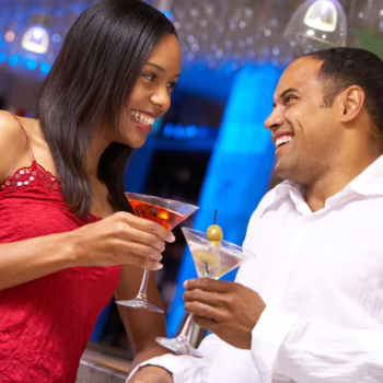 Smiling couple clinking cocktails and sharing a flirty moment at a bar, symbolizing trust, connection, and open communication in swinging relationships.