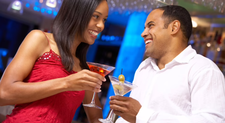 Smiling couple clinking cocktails and sharing a flirty moment at a bar, symbolizing trust, connection, and open communication in swinging relationships.