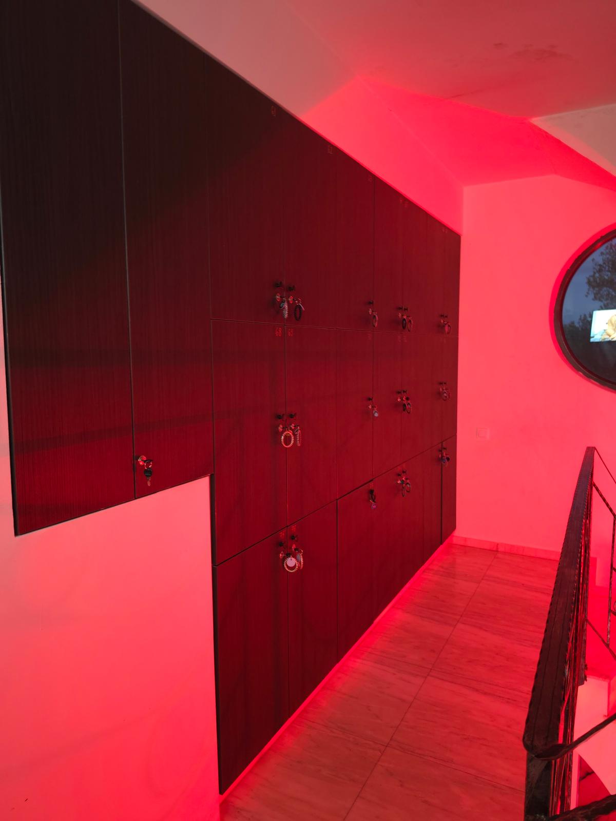 Dark wooden lockers with padlocks line a pink-lit hallway; circular window on the right reflects light.