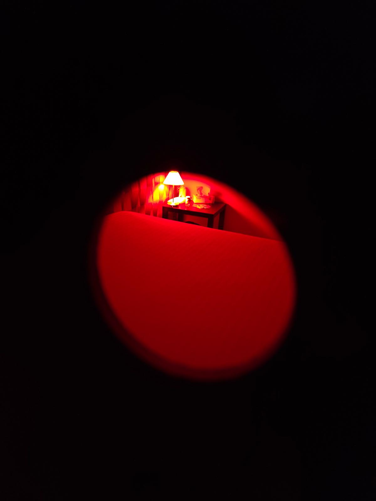Red-tinted night scene: a lit table lamp on a small bedside table beside a bed, viewed through a circular aperture in darkness