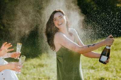 Woman laughing while opening a champagne bottle outdoors, symbolizing release, joy, and sexual awakening in a real erotic story about first time squirting.
