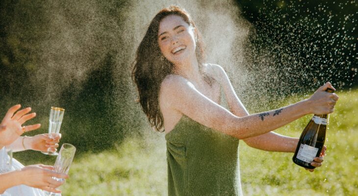 Woman laughing while opening a champagne bottle outdoors, symbolizing release, joy, and sexual awakening in a real erotic story about first time squirting.