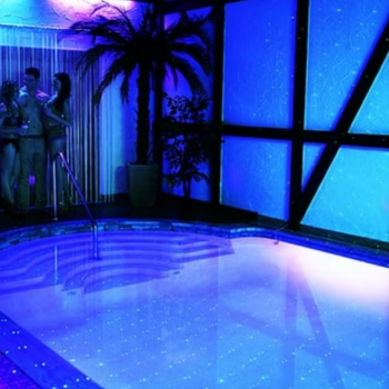 Indoor pool area with blue neon lighting and starry reflections, with a small group chatting near the curtained corner by the poolside.