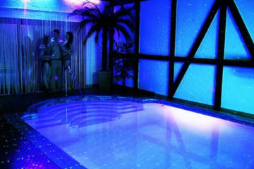 Indoor pool area with blue neon lighting and starry reflections, with a small group chatting near the curtained corner by the poolside.