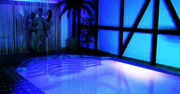 Indoor pool area with blue neon lighting and starry reflections, with a small group chatting near the curtained corner by the poolside.