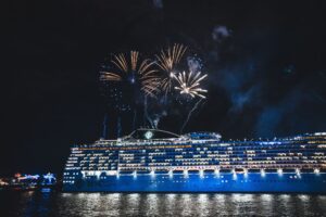 Large illuminated cruise ship at sea during a nighttime fireworks display with reflections on the water and a festive luxury travel atmosphere.
