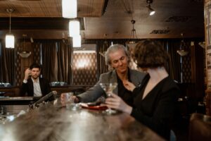 Man checking his phone while a couple enjoys drinks at a bar, illustrating anticipation and emotional distance in a hotwife relationship.